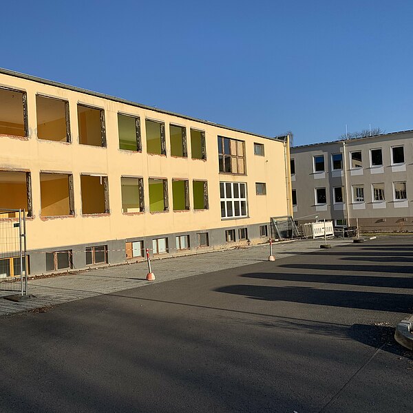 Zweistöckiges gelbes Schulgebäude im Rückbau ohne Fenster, Bauzaun und Absperrungen auf dem Hof, neben den beigefarbenen Neubau im Hintergrund, unter klarem blauem Himmel.