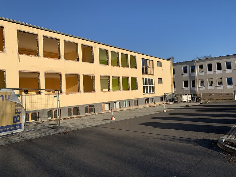 Zweistöckiges gelbes Schulgebäude im Rückbau ohne Fenster, Bauzaun und Absperrungen auf dem Hof, neben den beigefarbenen Neubau im Hintergrund, unter klarem blauem Himmel.